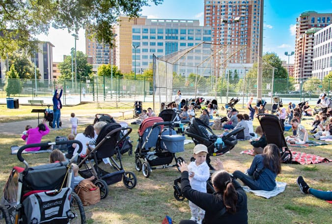 Families sitting in a park watching a speaker with city buildings in the distance